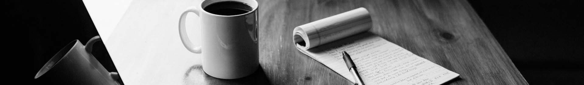 A photo of a coffee mug, pad of paper, and pen sitting on a desk.